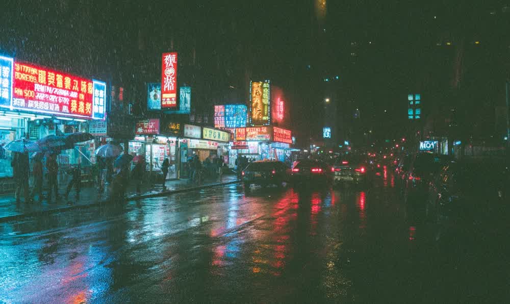 Neon signs reflected in rainy city pavement at night with storefront windows, umbrellas, and retro street style