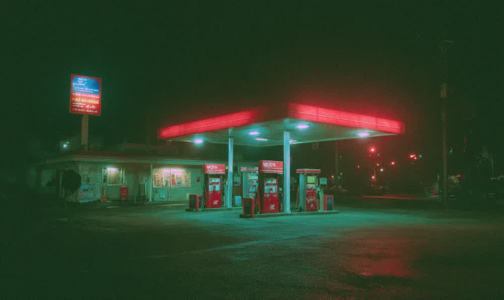 An old gas station sign glowing at night above wet pavement, vintage pumps, retro car lights, and a dark roadside