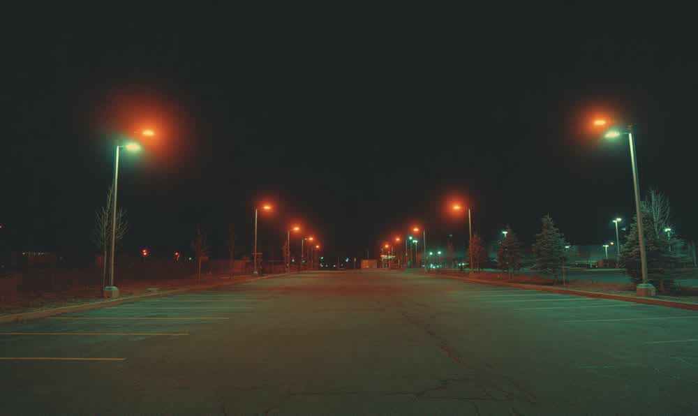 An empty retro parking lot under streetlights at night with wet asphalt, distant storefronts, and a lone parked car