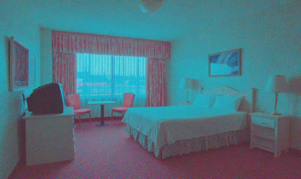 A retro motel room with patterned bedspread, glowing sign outside, old TV, wall art, luggage, and parking lot light