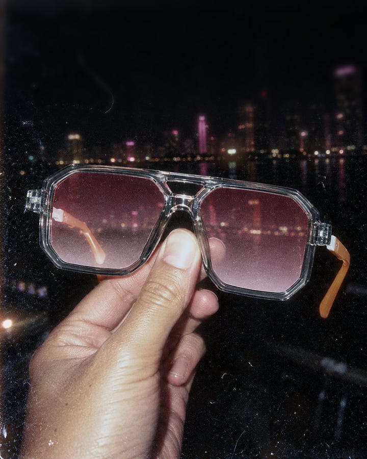 Pink Haze Sunglasses 1984 held up against a night city skyline